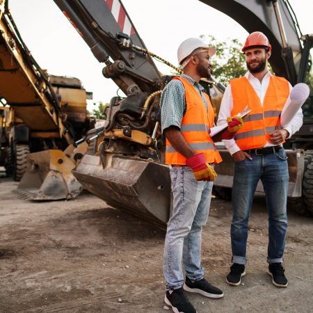Two men engineers in workwear discussing their work standing against construction machines